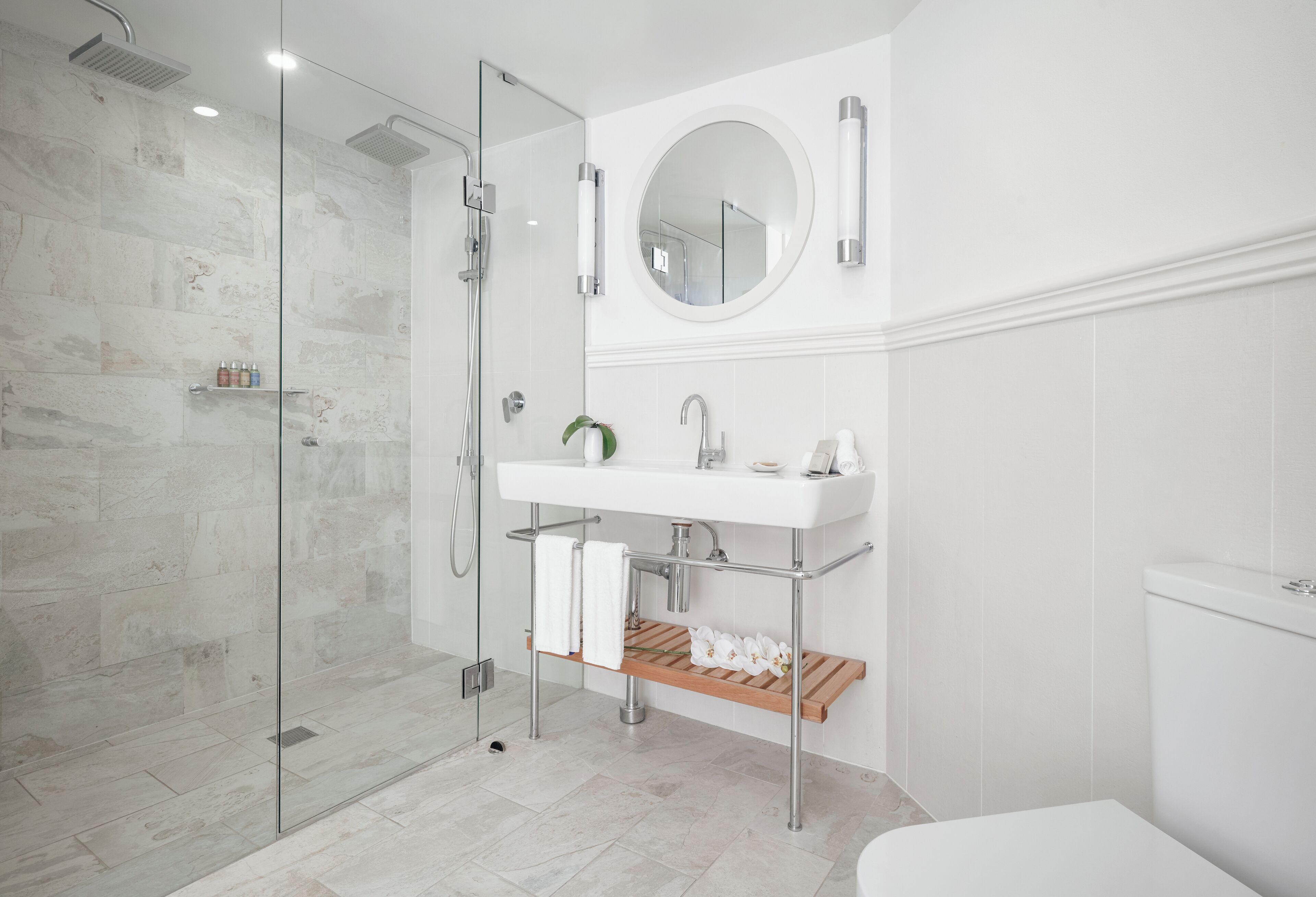 Ein modernes Badezimmer mit einer ein Glasfach raumseitig geschlossenen Dusche, einem weißen Waschbecken mit Spiegel darüber und einer Holzablage mit Handtüchern und einer Topfpflanze.