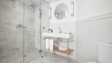 Ein modernes Badezimmer mit einer ein Glasfach raumseitig geschlossenen Dusche, einem weißen Waschbecken mit Spiegel darüber und einer Holzablage mit Handtüchern und einer Topfpflanze.