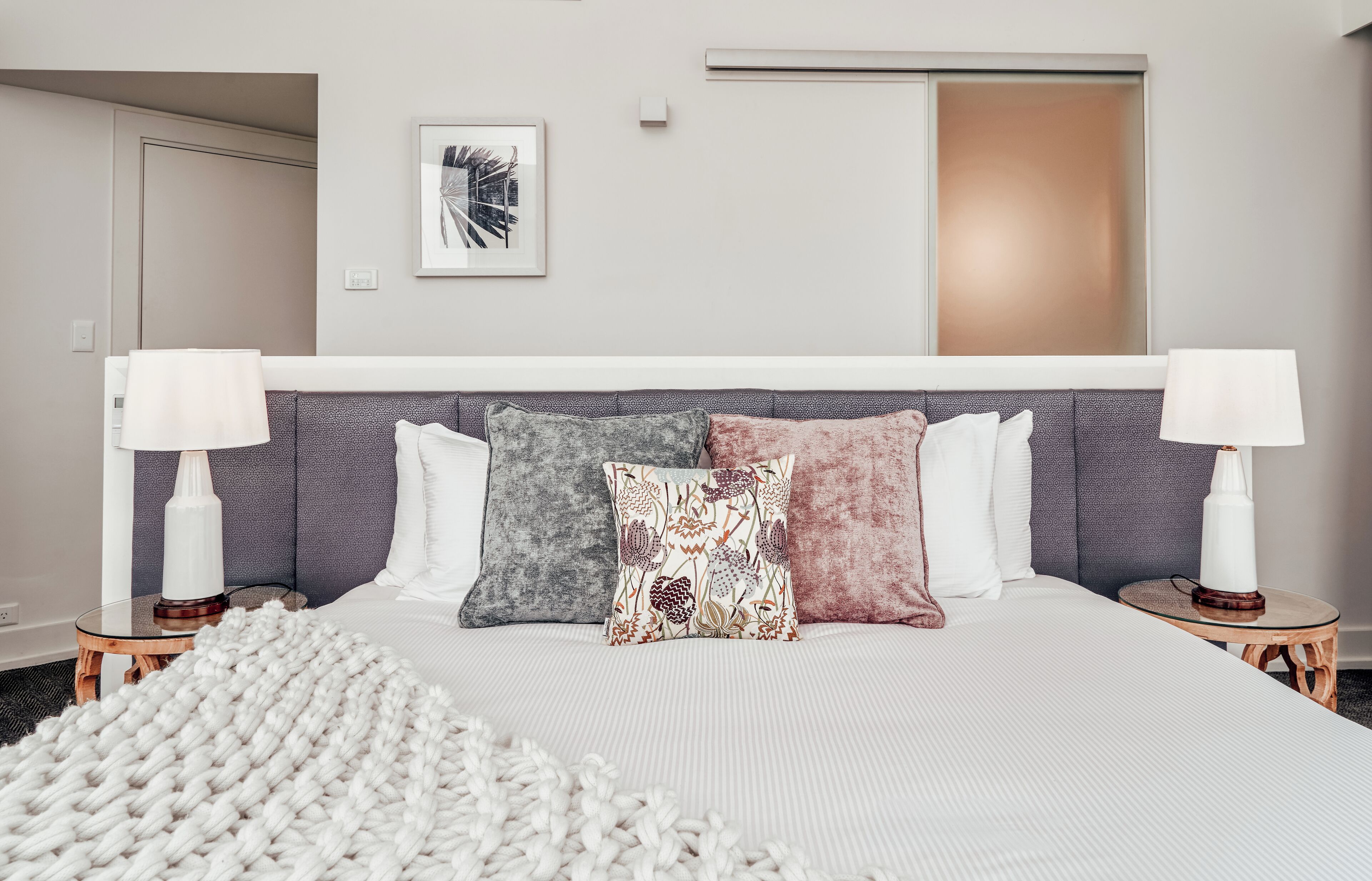 A hotel room with a large bed, two bedside tables with lamps, a gray headboard, and a framed picture on the wall.