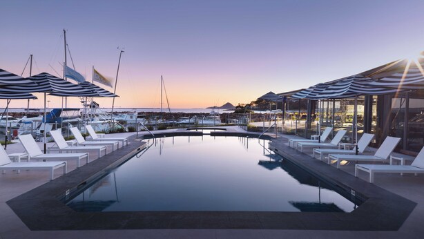 A swimming pool with lounge chairs and umbrellas, surrounded by boats and a scenic view. - Anchorage Port Stephens (Corlette)