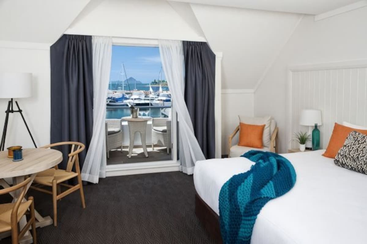A hotel room with a bed, a desk, a chair, a small table, a window with curtains, and a view of boats.