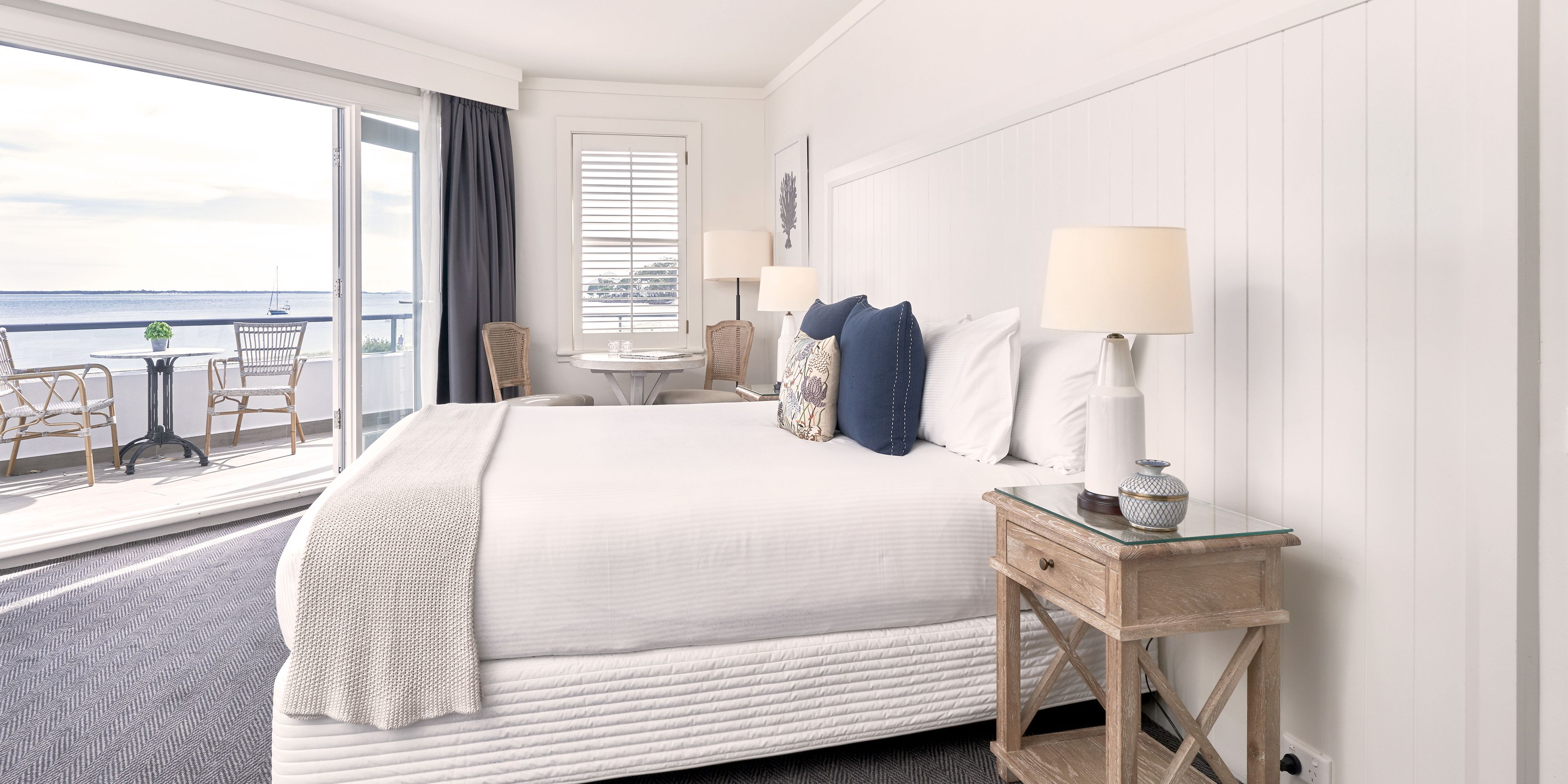 A hotel room with a large bed, a glass table, two chairs, a window with a view of the sea, and a balcony with a table and chairs.