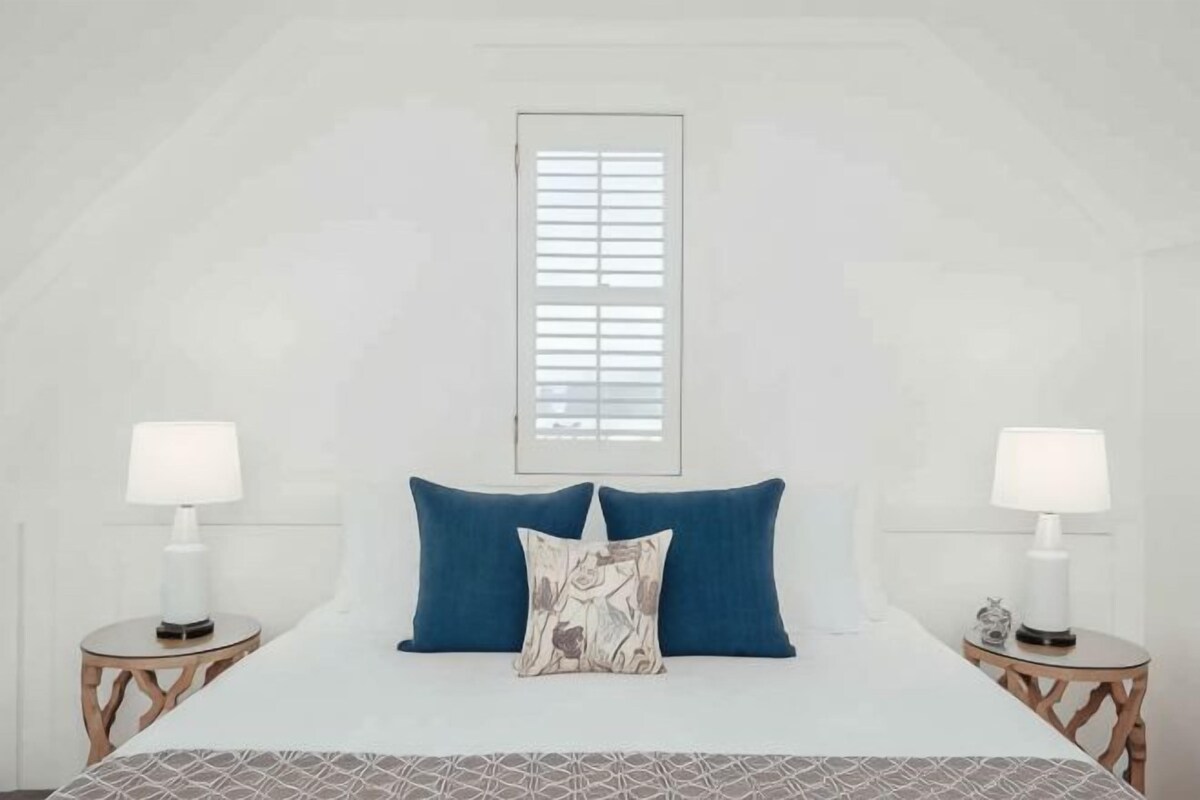 A bedroom with a bed, two bedside tables, a window with blinds, and a lamp on each table.
