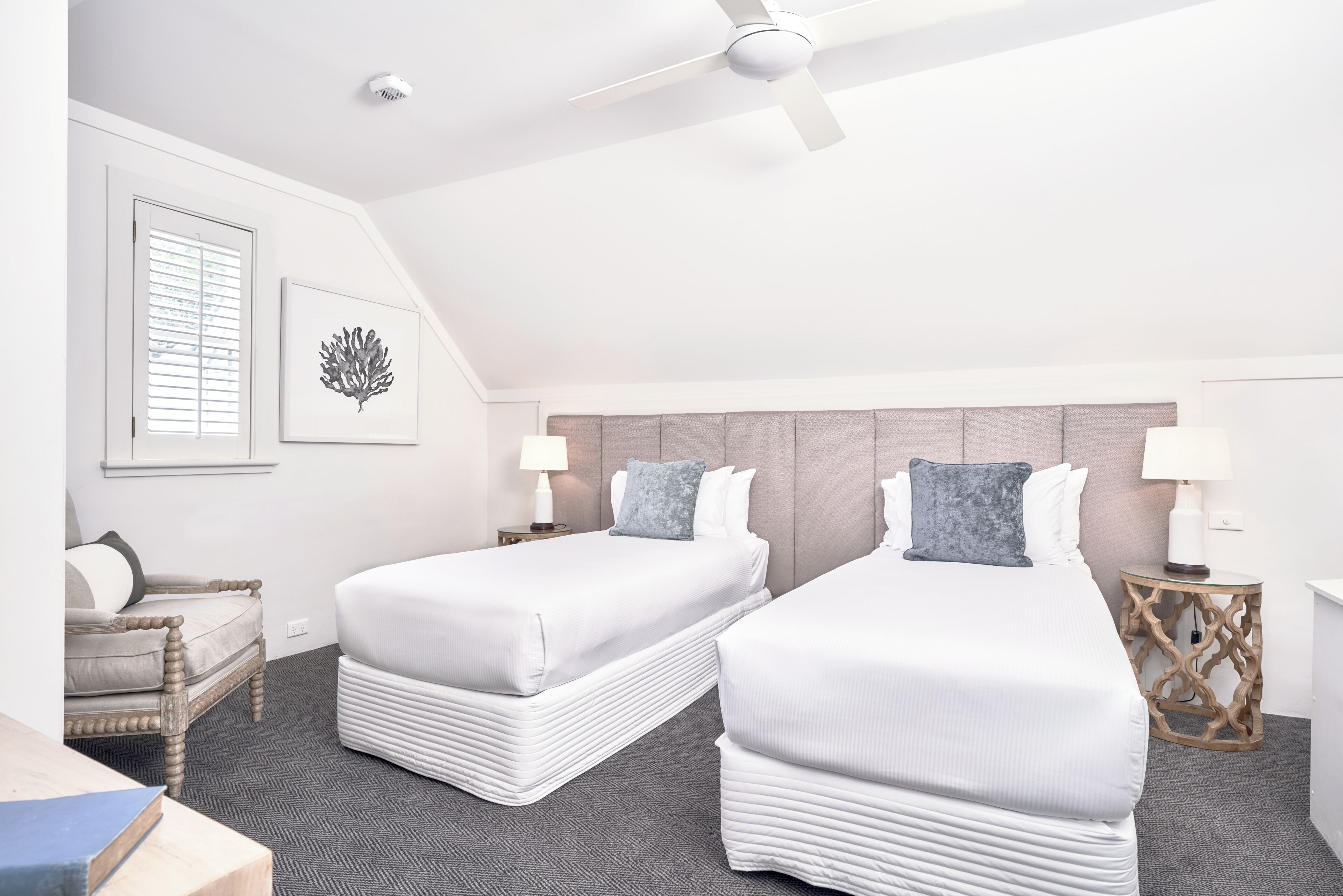 Habitación de hotel con dos camas, un ventilador de techo, una ventana con persianas, una obra de arte en la pared y una mesita de noche con lámpara.