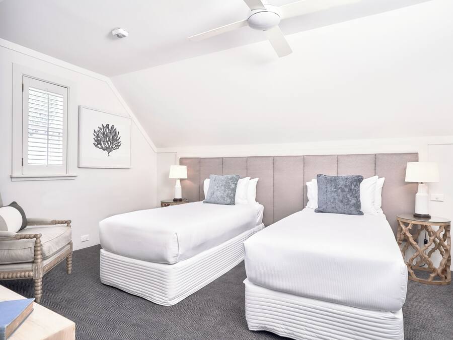 Habitación de hotel con dos camas, un ventilador de techo, una ventana con persianas, una obra de arte en la pared y una mesita de noche con lámpara.