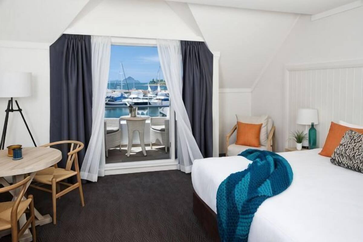 A hotel room with a bed, a desk, a chair, a small table, a window with curtains, and a view of boats.