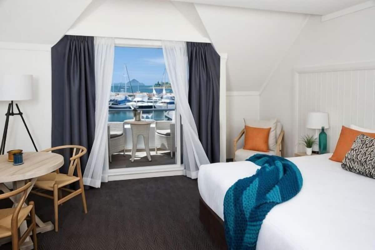 A hotel room with a bed, a desk, a chair, a small table, a window with curtains, and a view of boats.