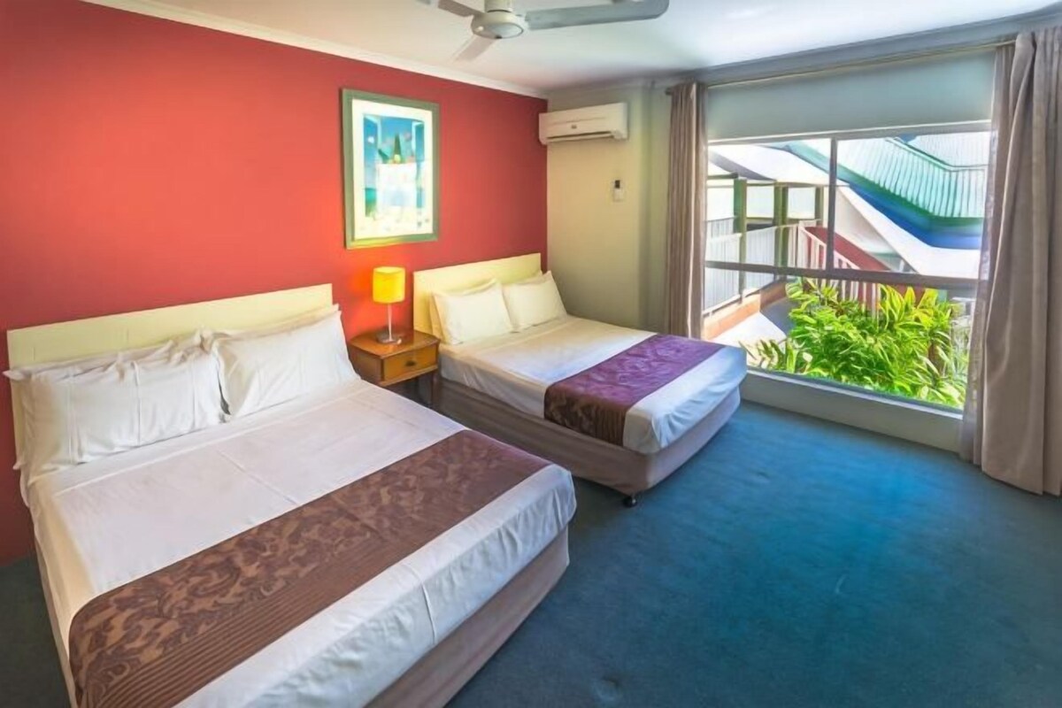 Een hotelkamer met twee bedden, een plafondventilator, een raam met uitzicht op een gebouw en groen, plus een airconditioning.