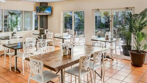 Un comedor amplio con sillas y mesas blancas, ventanas grandes y plantas en macetas.