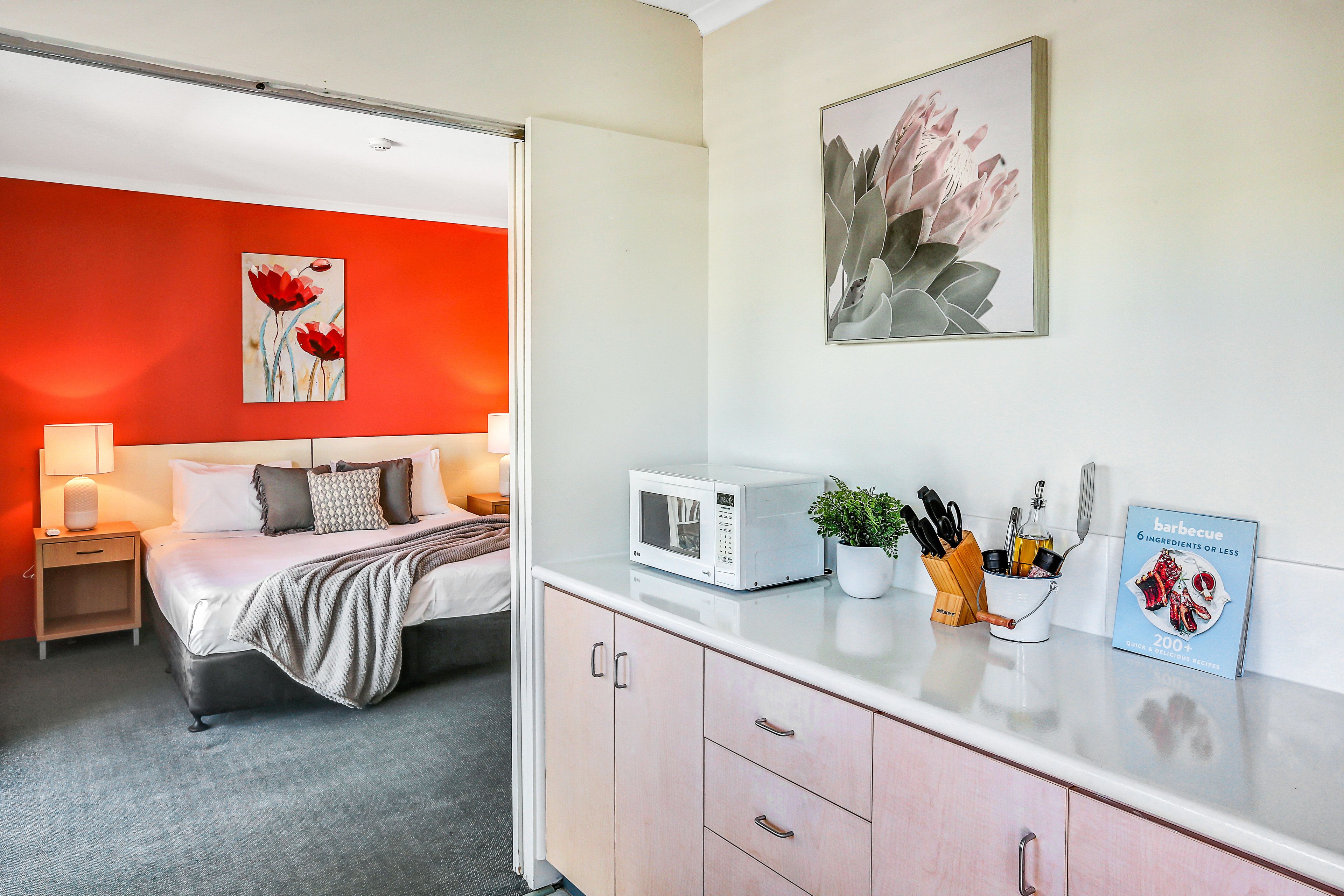 A modern hotel room with a bed, bedside table, and a kitchenette area with a microwave, a potted plant, and a cookbook.