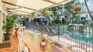 Un espacio al aire libre techado con piscina, barra de madera, sillas blancas y plantas en macetas.
