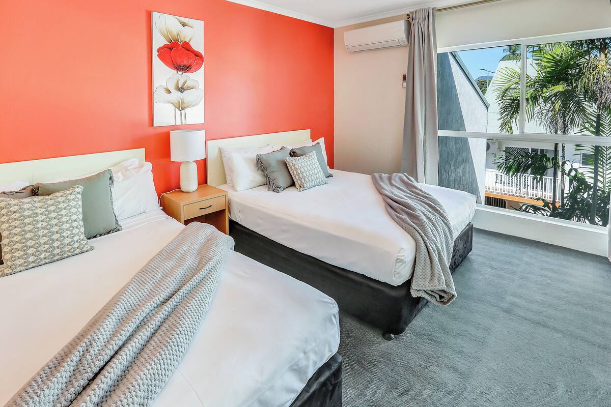 A hotel room with two beds, a nightstand, a lamp, a large window with a view of palm trees, and a red accent wall.