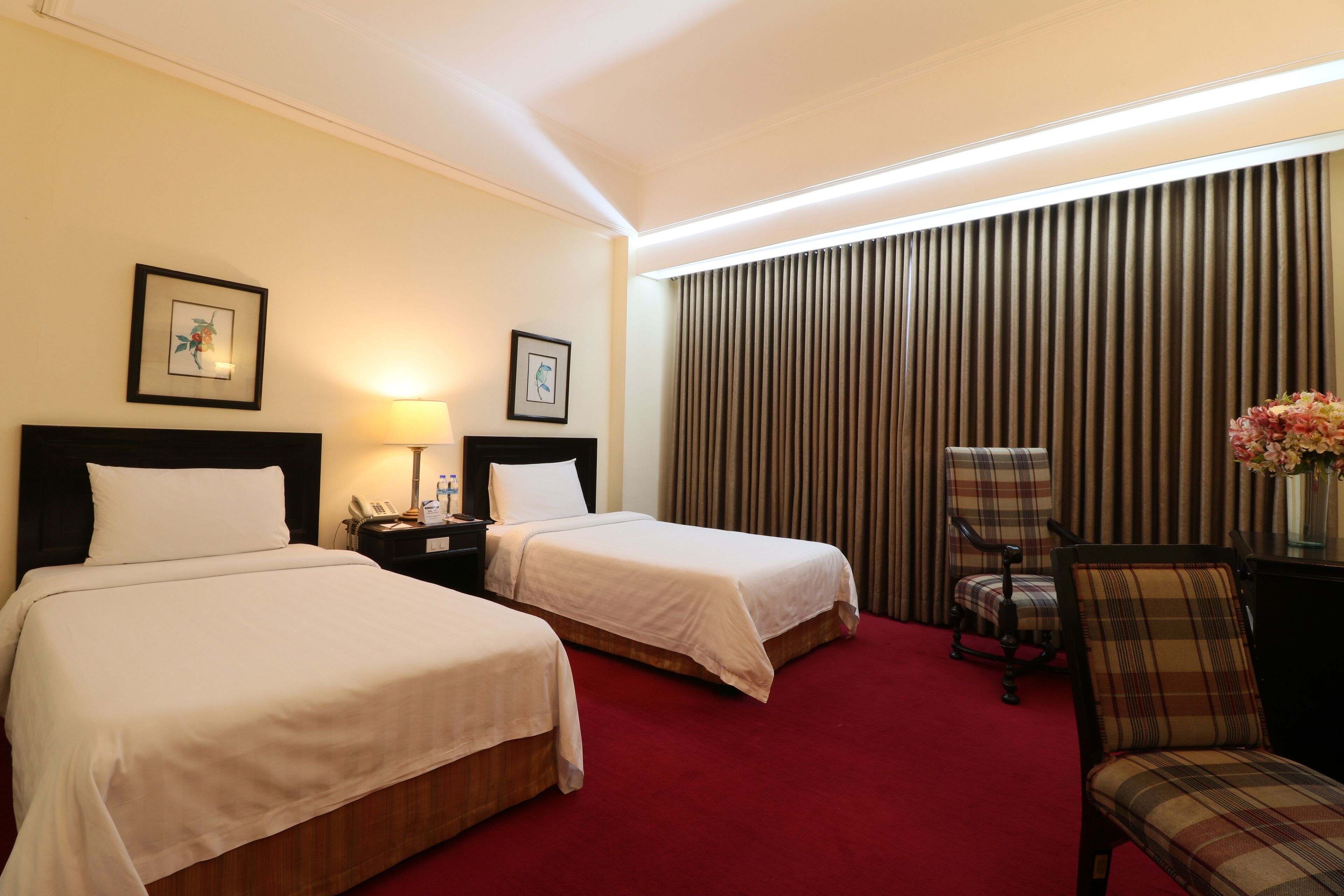 A hotel room with two beds, a desk with a lamp, a chair, and a large window with curtains.