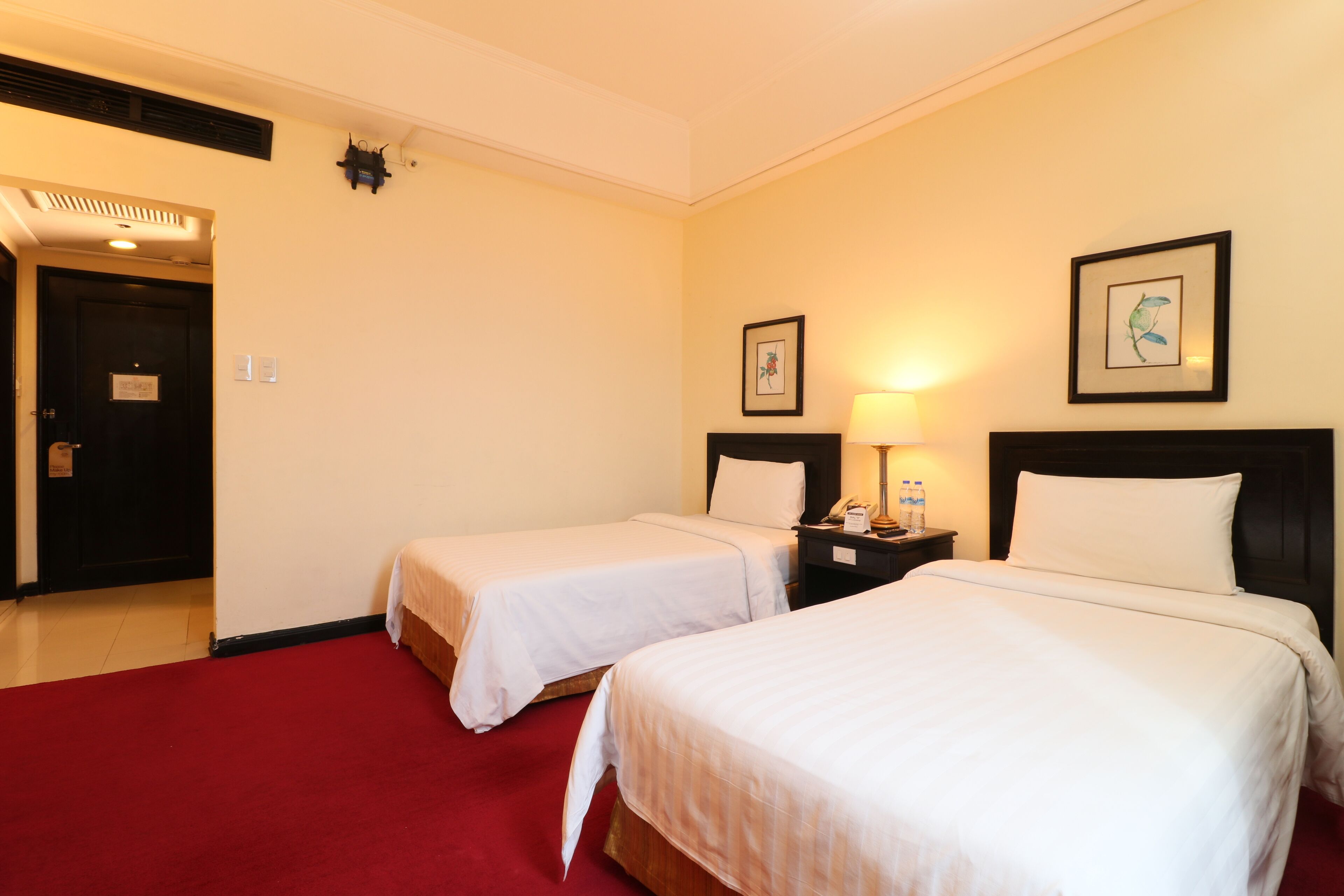 Quarto de hotel com duas camas de solteiro, um tapete vermelho, mesa de cabeceira com abajur e uma porta com placa.