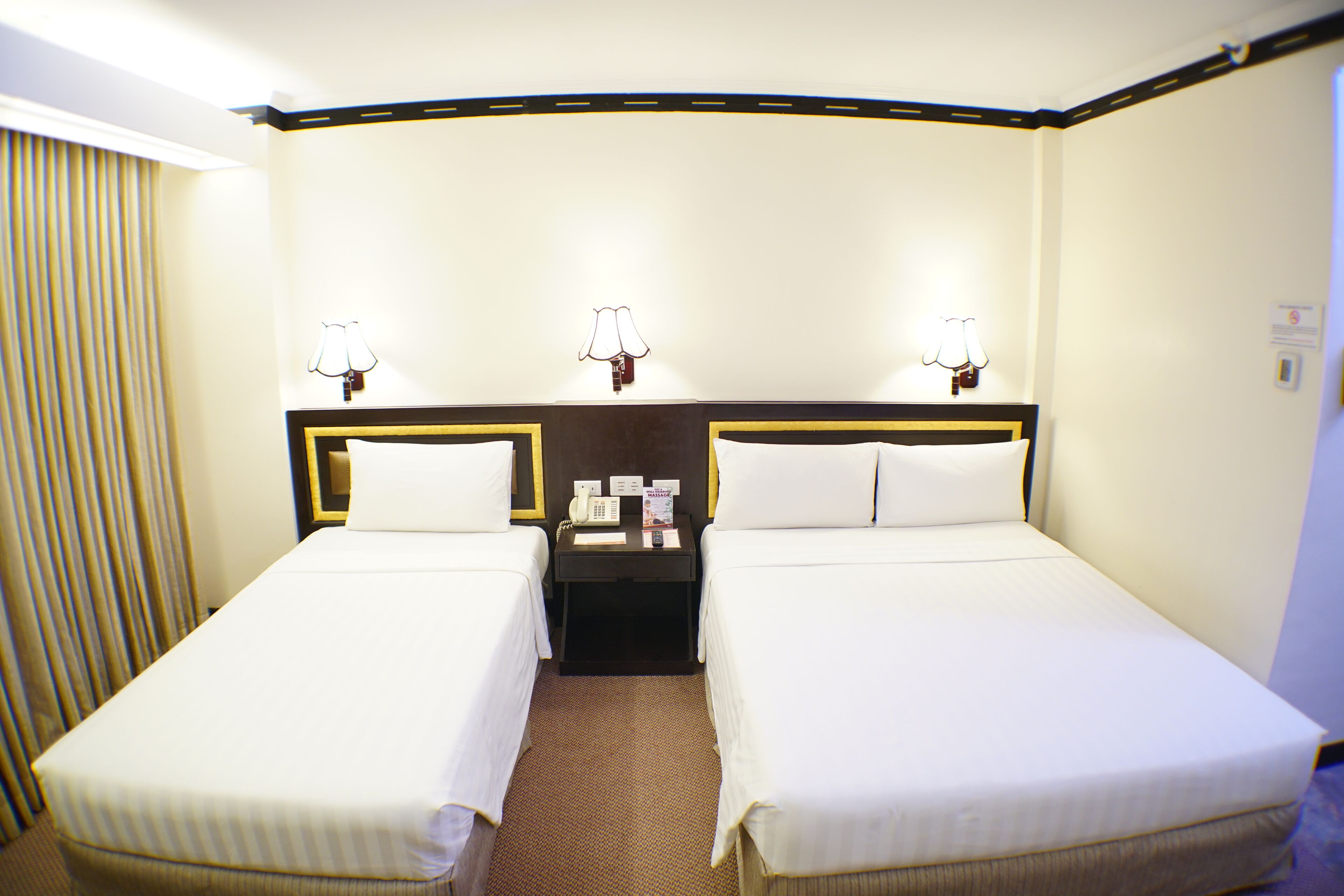 A hotel room with two single beds, a small table with a telephone and a lamp, and a curtain on the left side.