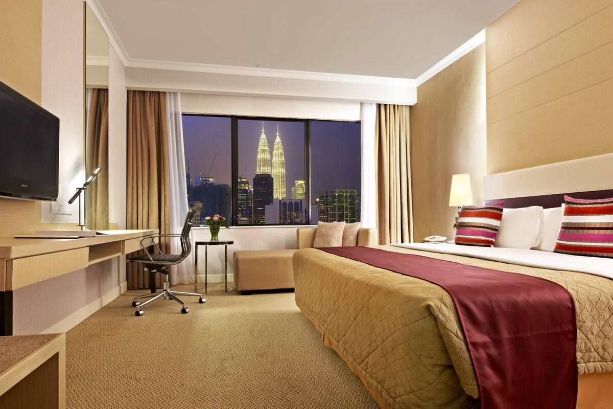 A hotel room with a bed, desk, chair, and a large window with a city view.