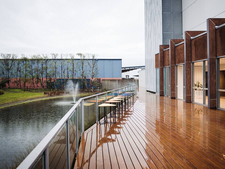 Una terraza de madera con un elemento acuático, una fila de mesas y un edificio moderno con ventanas amplias.