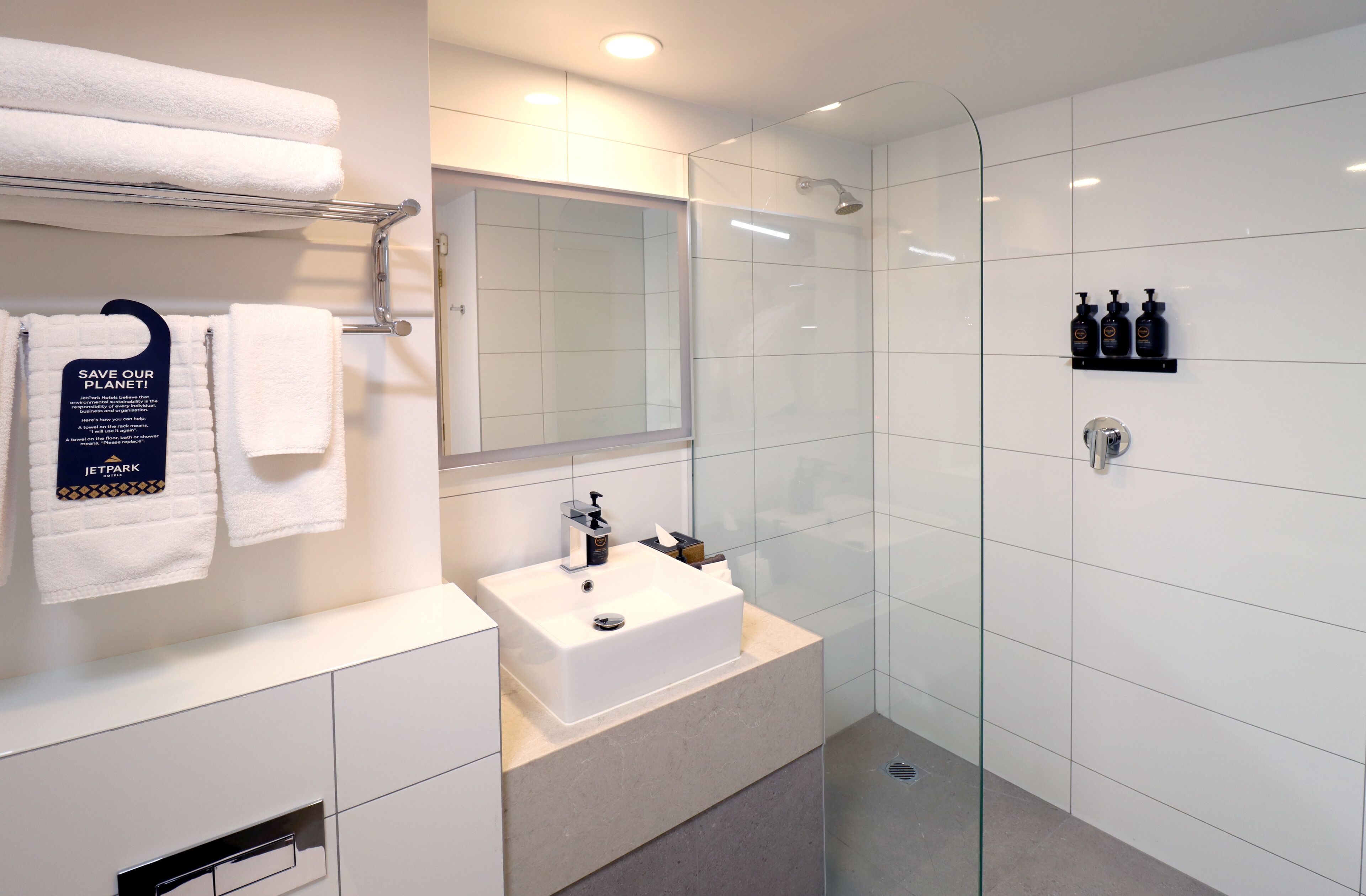 A modern bathroom with a glass shower enclosure, a white sink, and a towel rack with a 'SAVE OUR PLANET' sign.