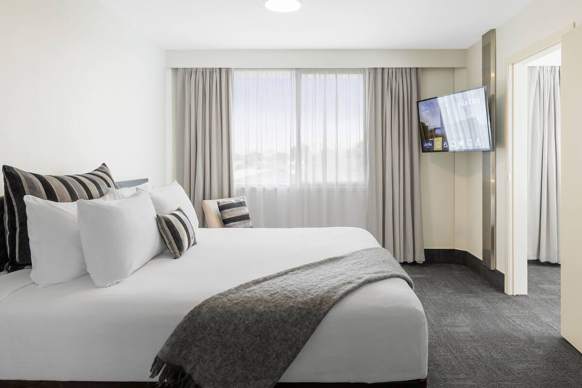 A hotel room with a large bed, a flat-screen TV, and a view of the outdoors through sheer curtains.
