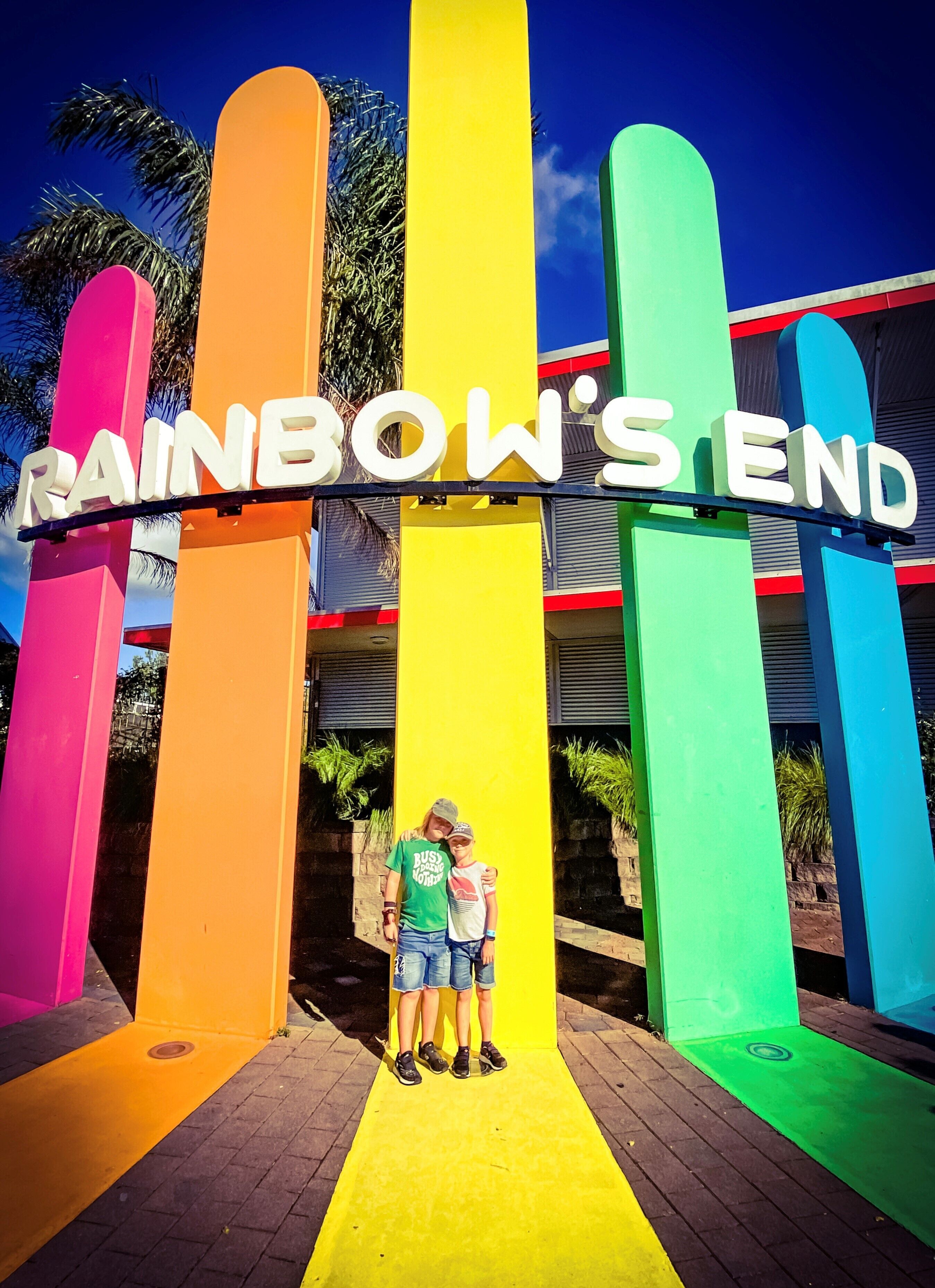 Ein farbiges Schild mit dem Schriftzug "RAINBOW'S END" und zwei Personen davor.