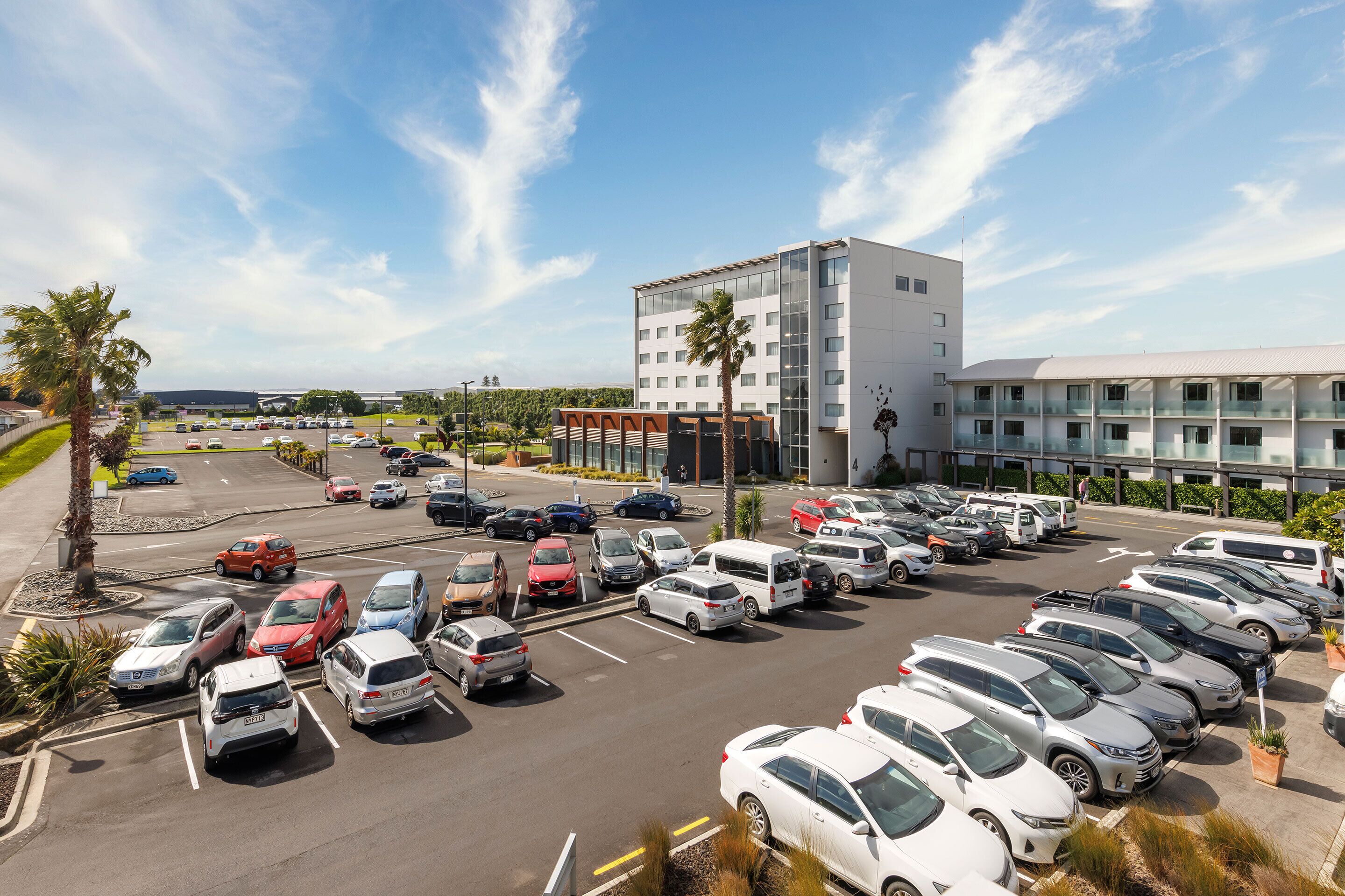 A parking lot with multiple rows of parked cars, a modern building with glass windows, and palm trees.