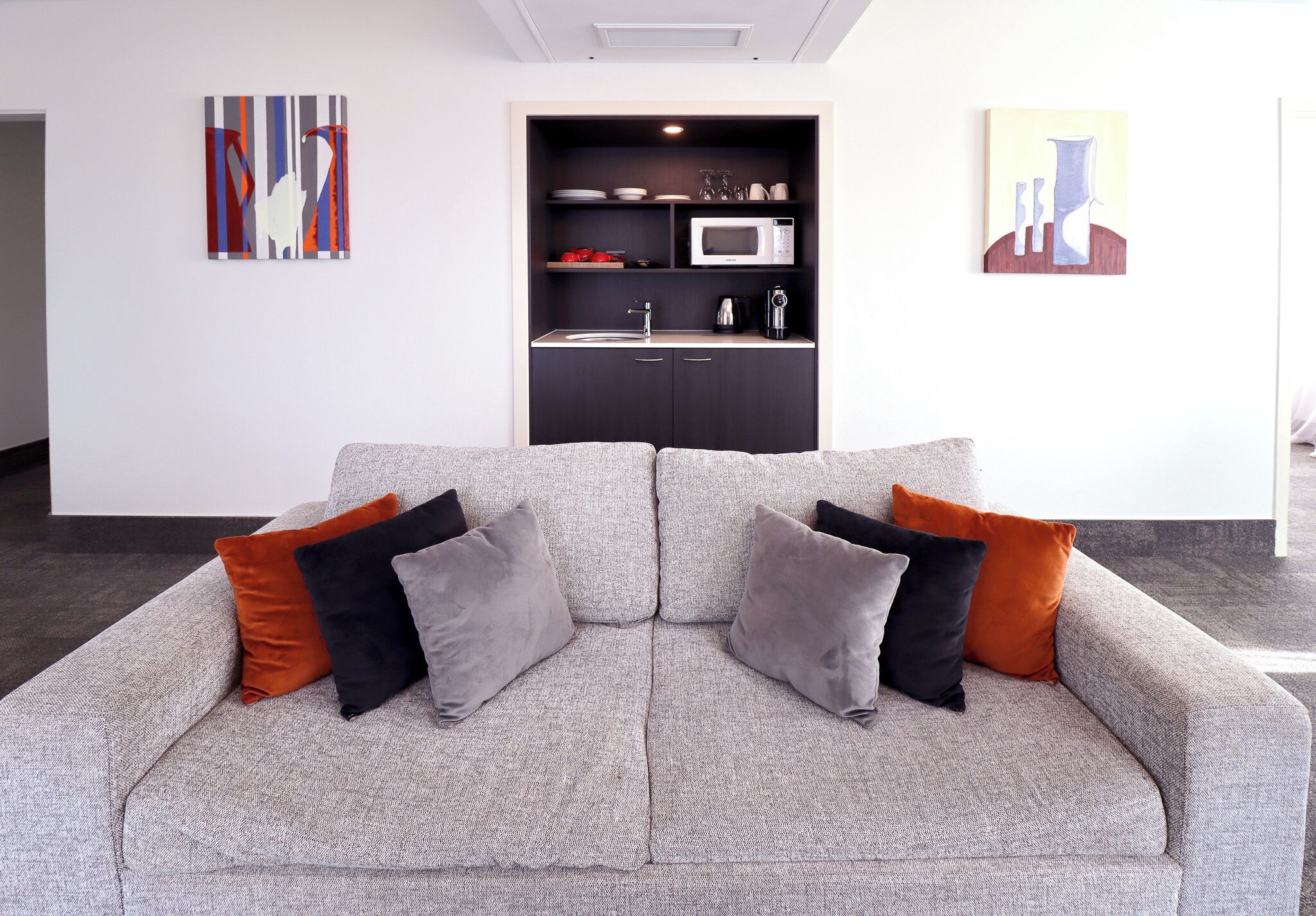 A modern living room with a grey sofa, black and orange cushions, a kitchen area with a microwave, and artwork on the walls.