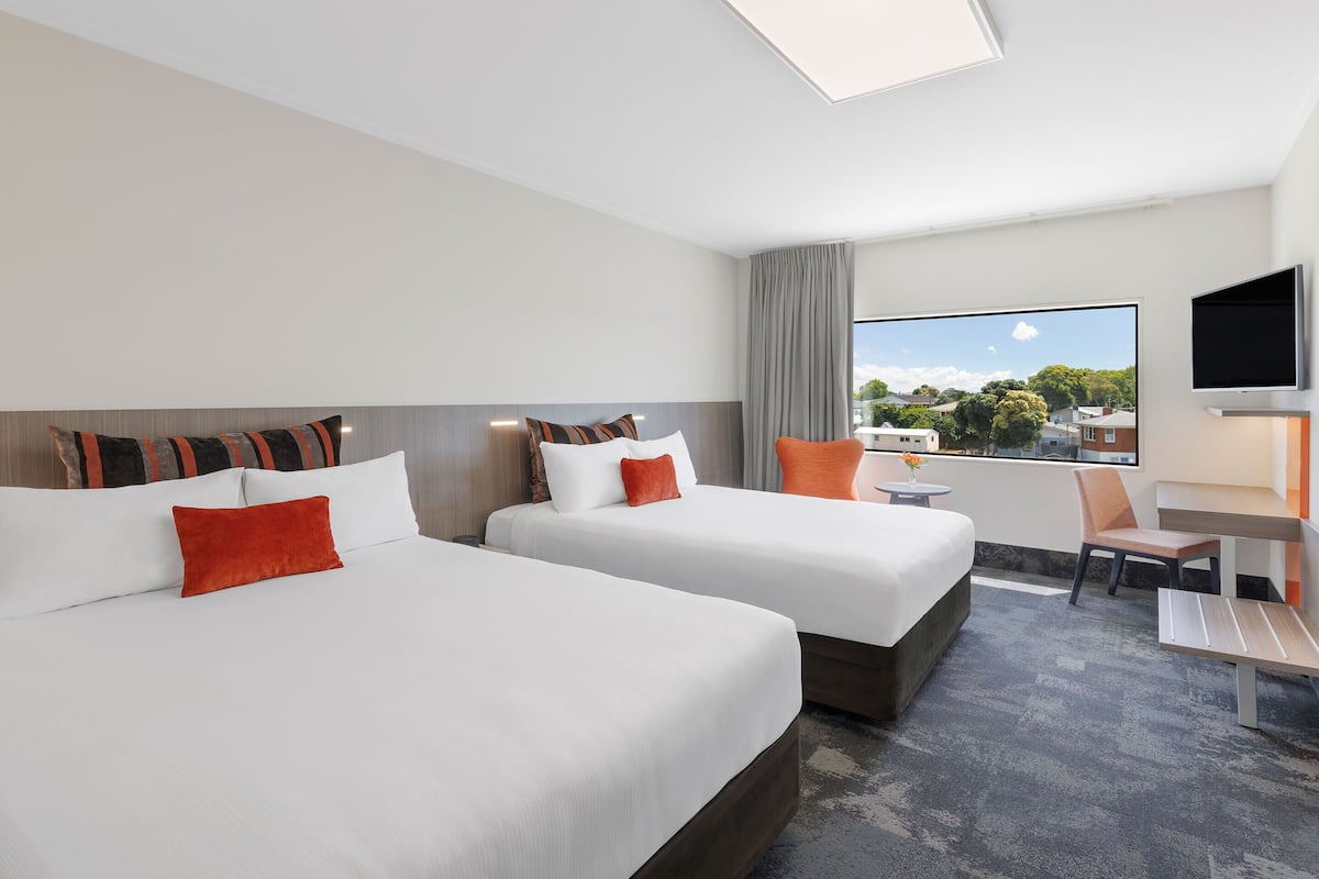 A hotel room with two beds, a TV, a desk, and a window with a city view.