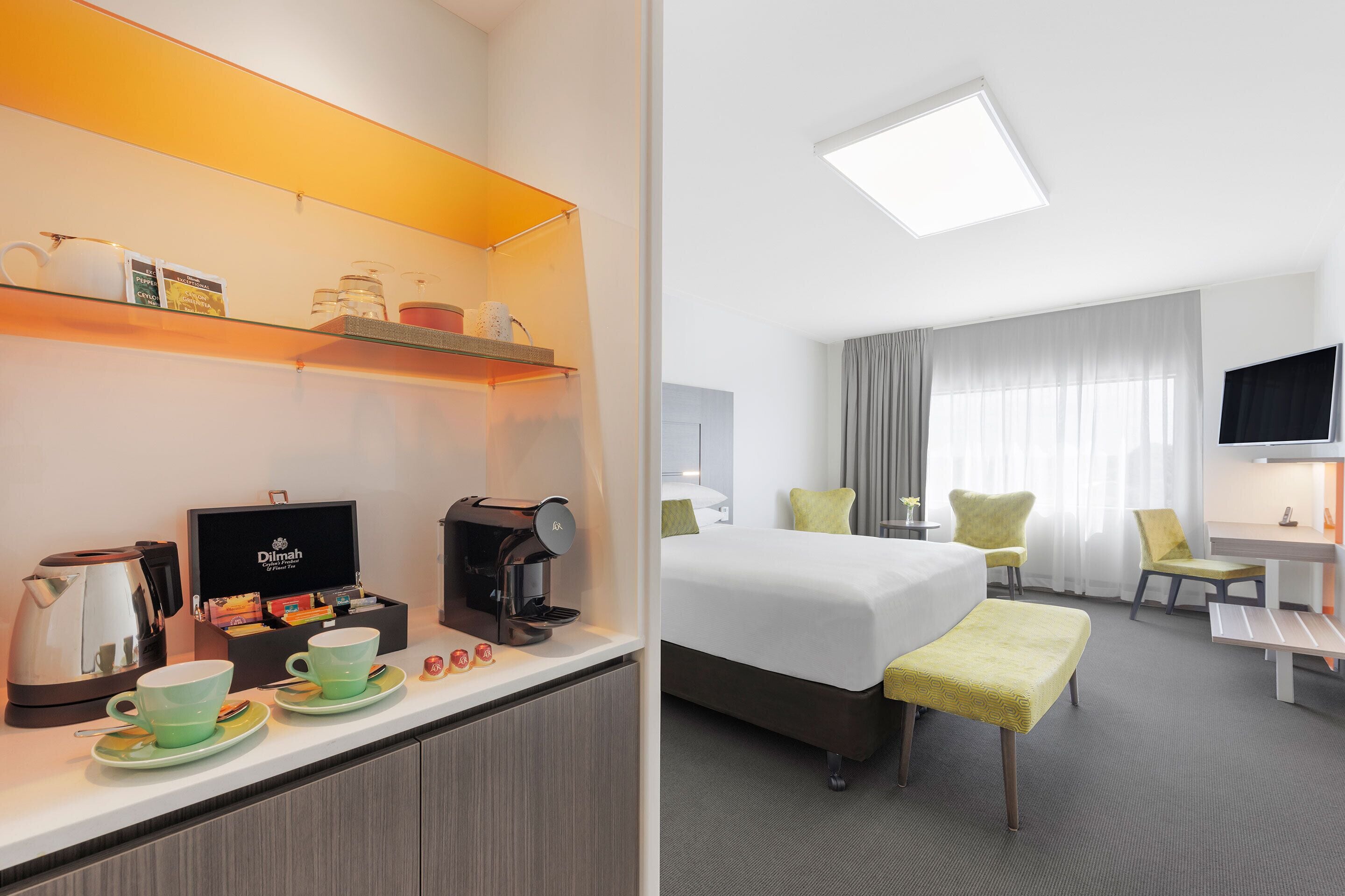 A hotel room with a bed, a desk, a chair, a coffee machine, and a shelf with cups and a box.