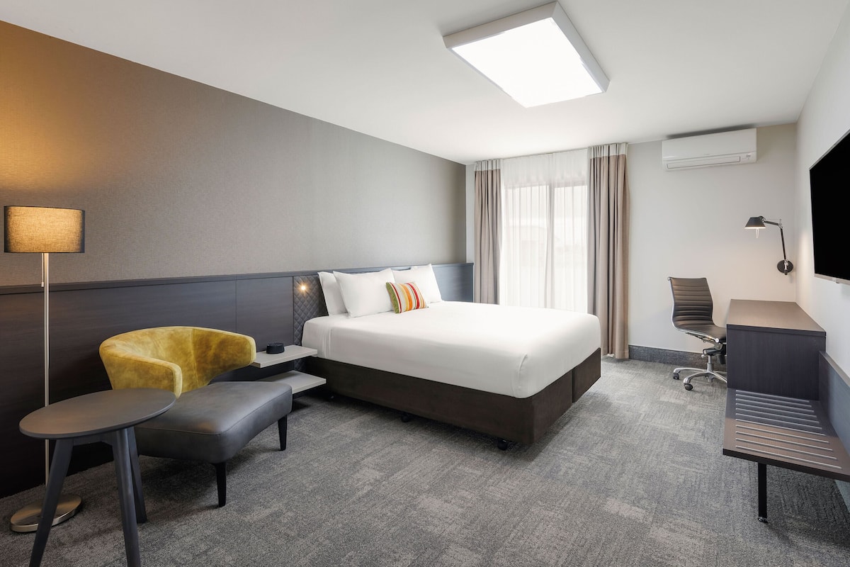 A hotel room with a bed, a desk, a chair, a small table, and a TV.
