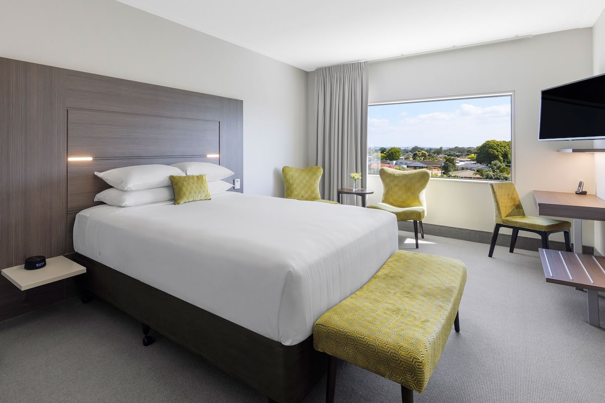 A hotel room with a large bed, a desk, two chairs, a bench, a small table with a vase, and a TV mounted on the wall.
