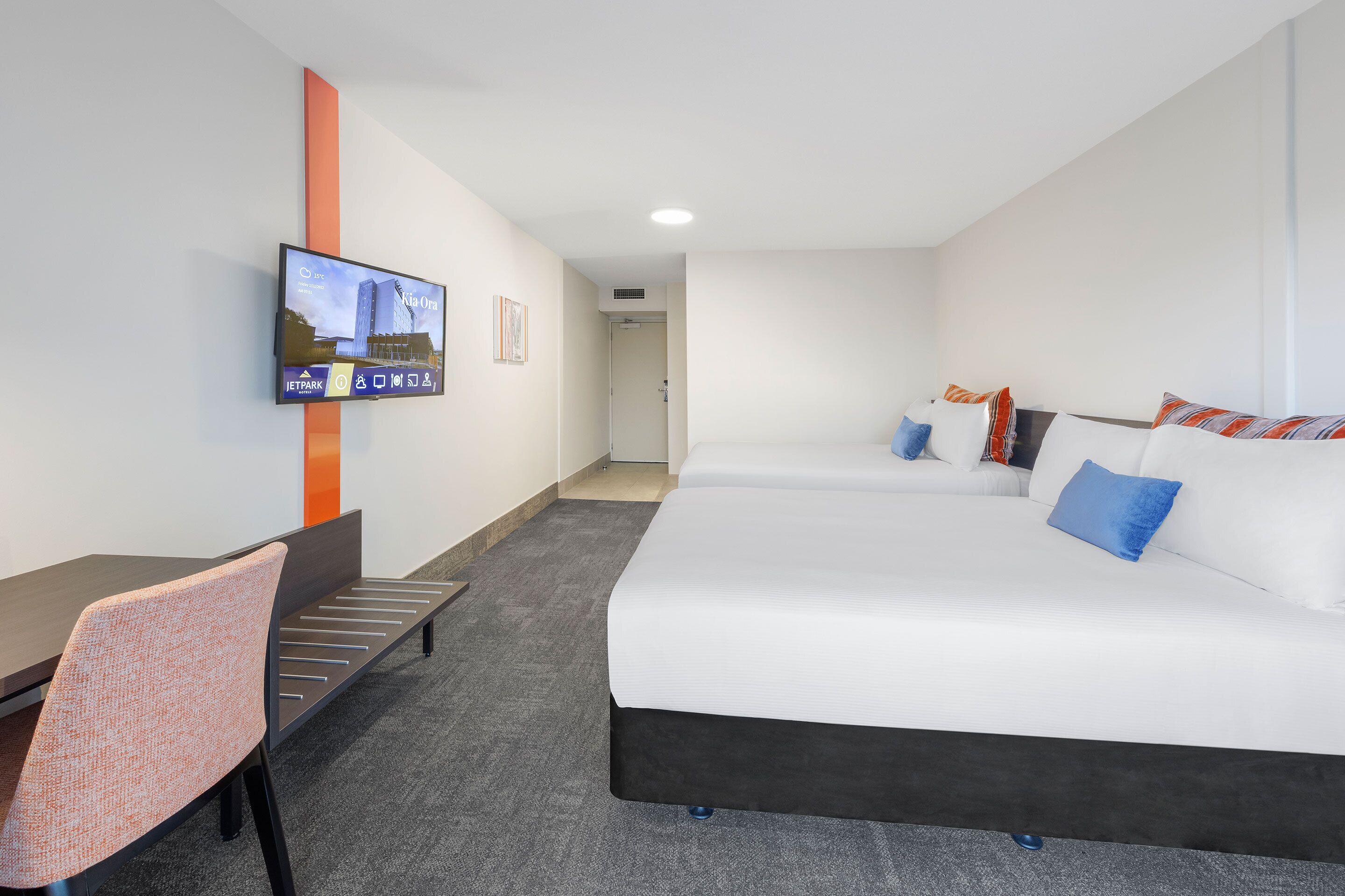 A hotel room with a bed, a desk, a chair, a TV mounted on the wall, and a door leading to another room.