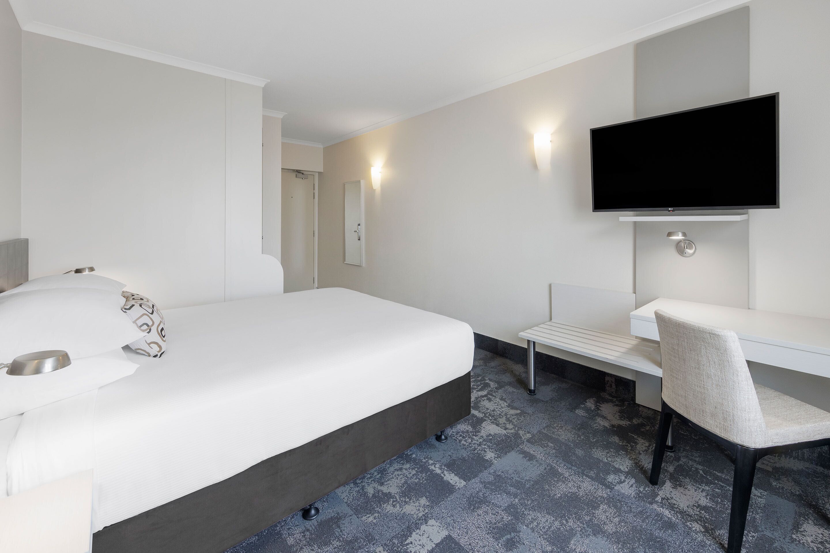 Une chambre d’hôtel avec un lit, un bureau, une chaise, une télévision et une salle de bain visible au fond.