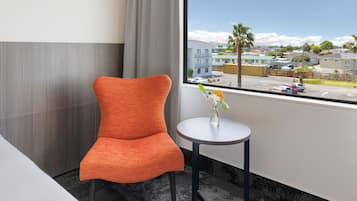 A hotel room with a modern orange chair, a small round table with a vase of flowers, and a large window offering a view of the street and buildings.