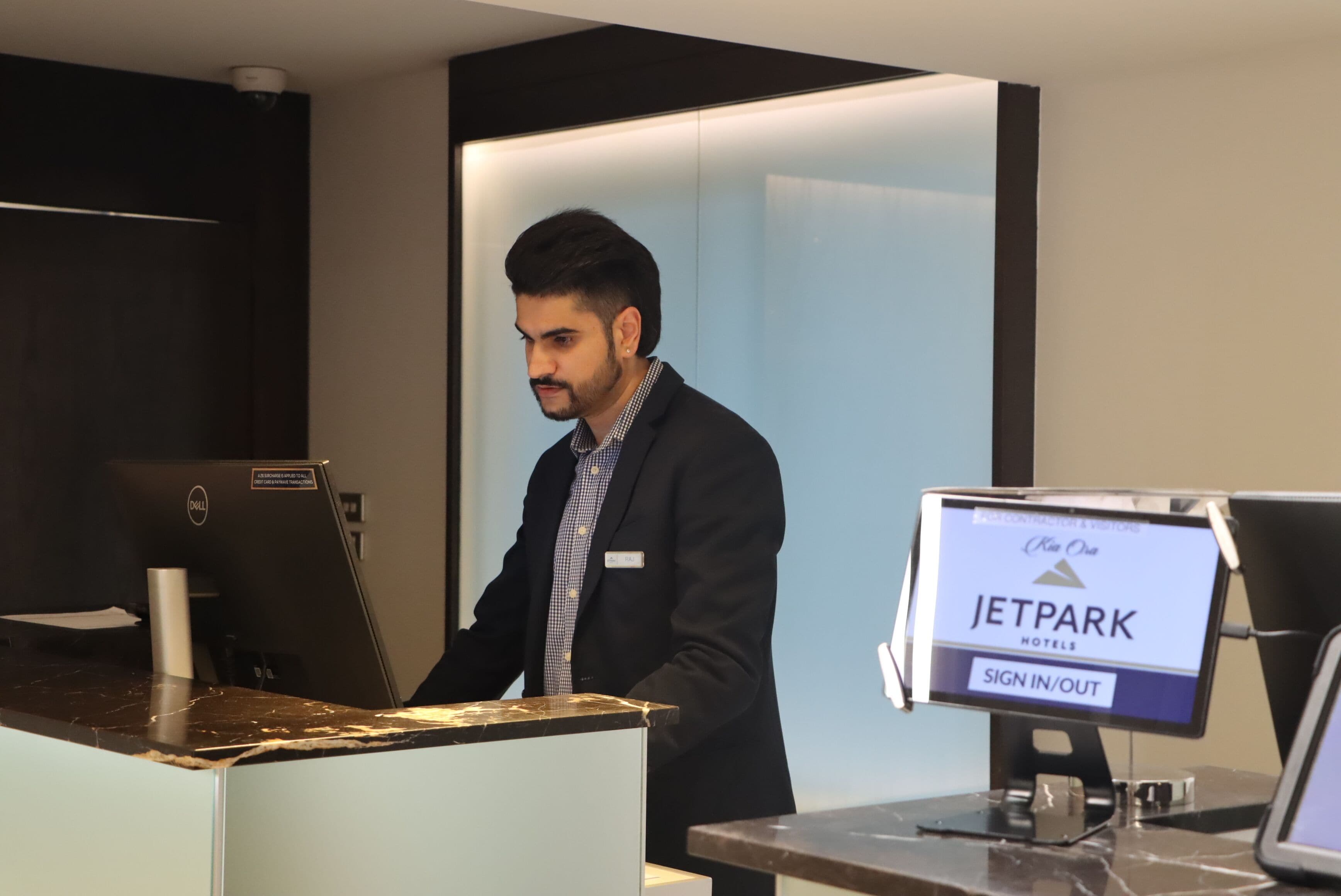 A man at a JetPark hotel reception desk with a sign-in/out monitor.