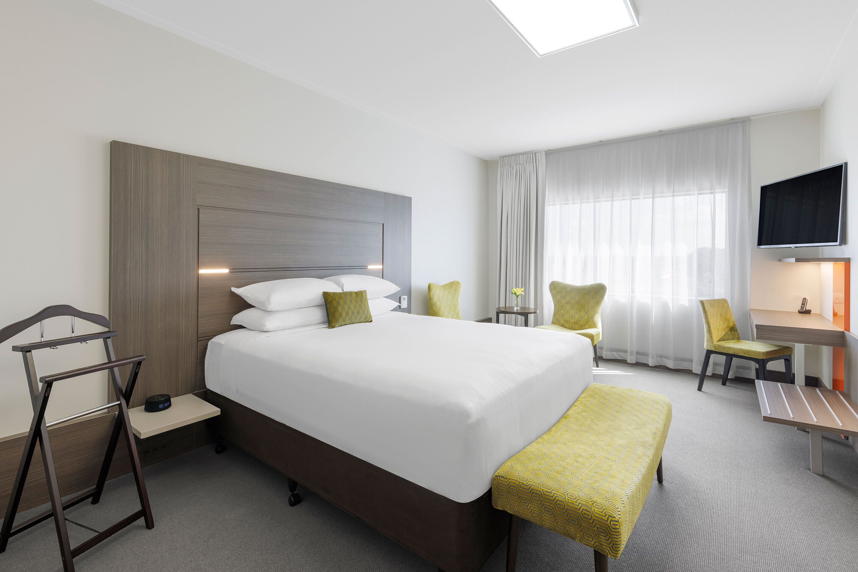 A hotel room with a large bed, bedside tables, a desk, a chair, a TV, and a window with curtains.