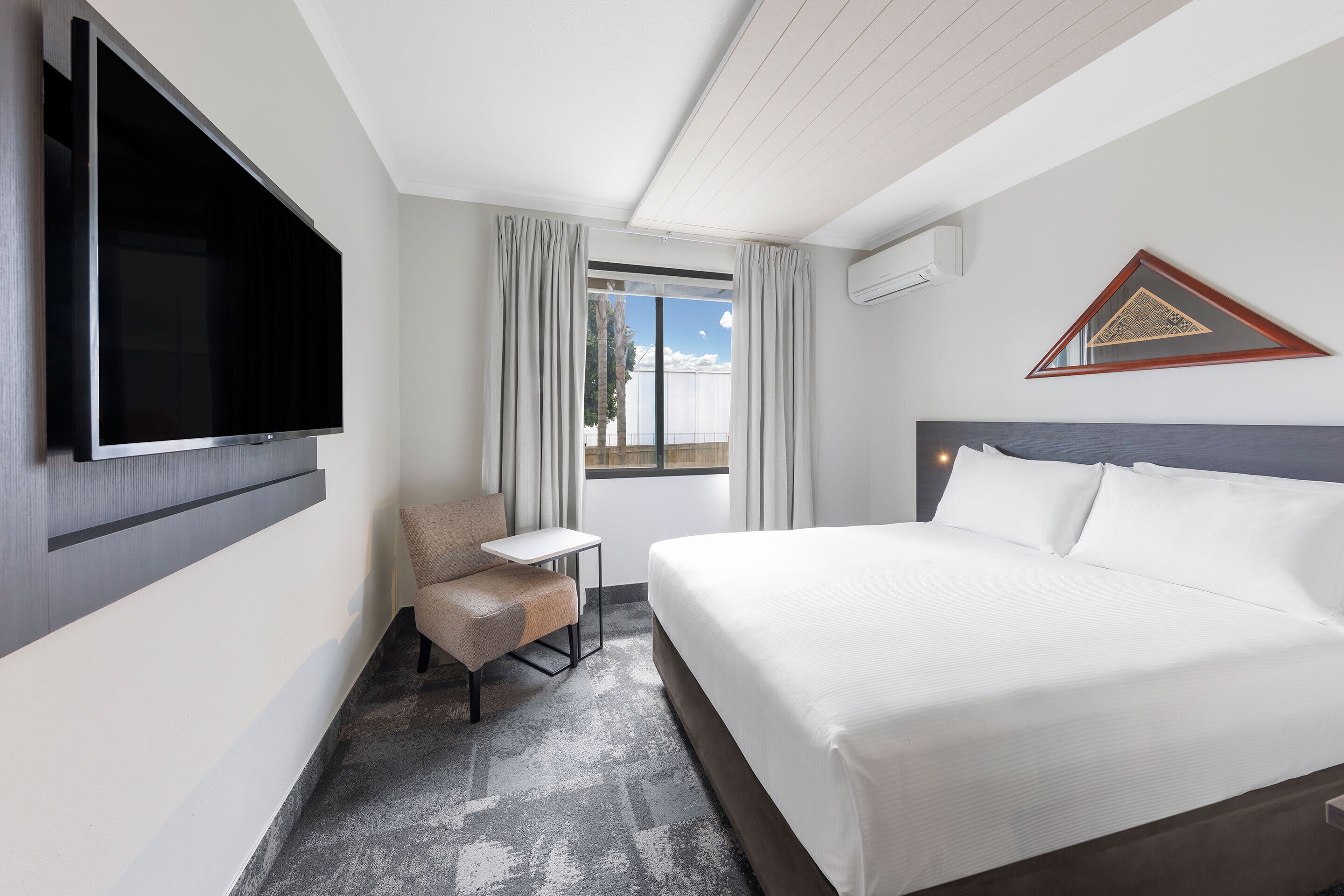 A hotel room with a bed, a TV mounted on the wall, a small table, a chair, and a window with curtains.