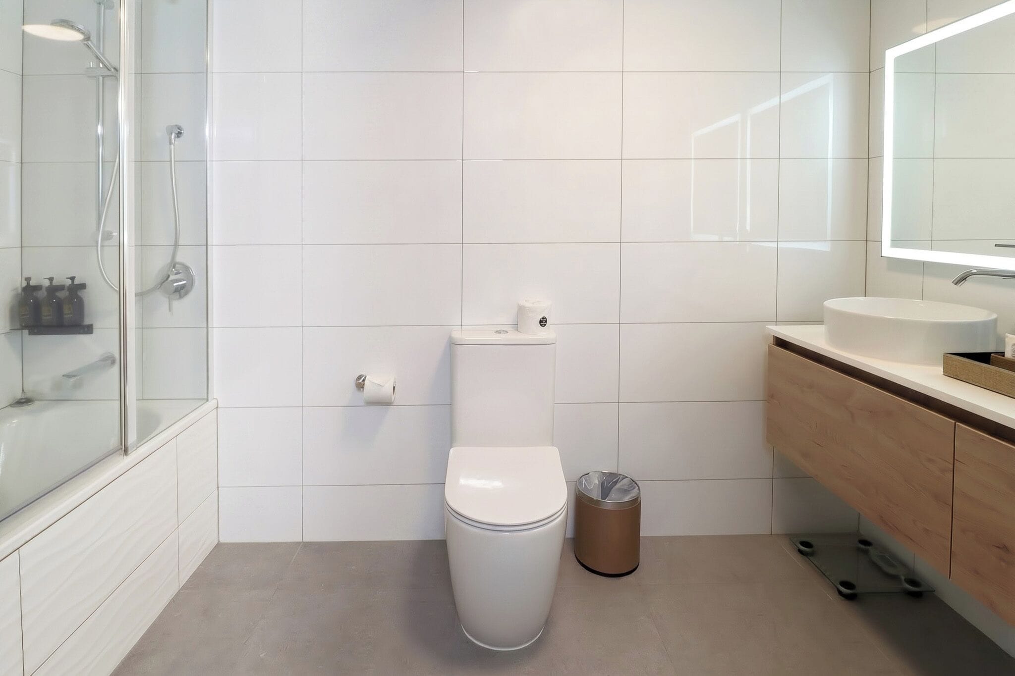 A modern bathroom with a toilet, a trash bin, a shower, and a vanity with a mirror.