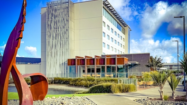 A modern hotel with a glass facade, a rooftop pool, and a distinctive metal sculpture.