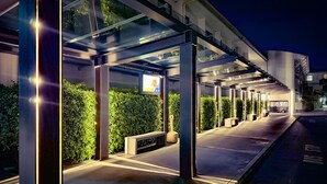 A modern hotel entrance with a covered walkway, greenery, and a television screen.