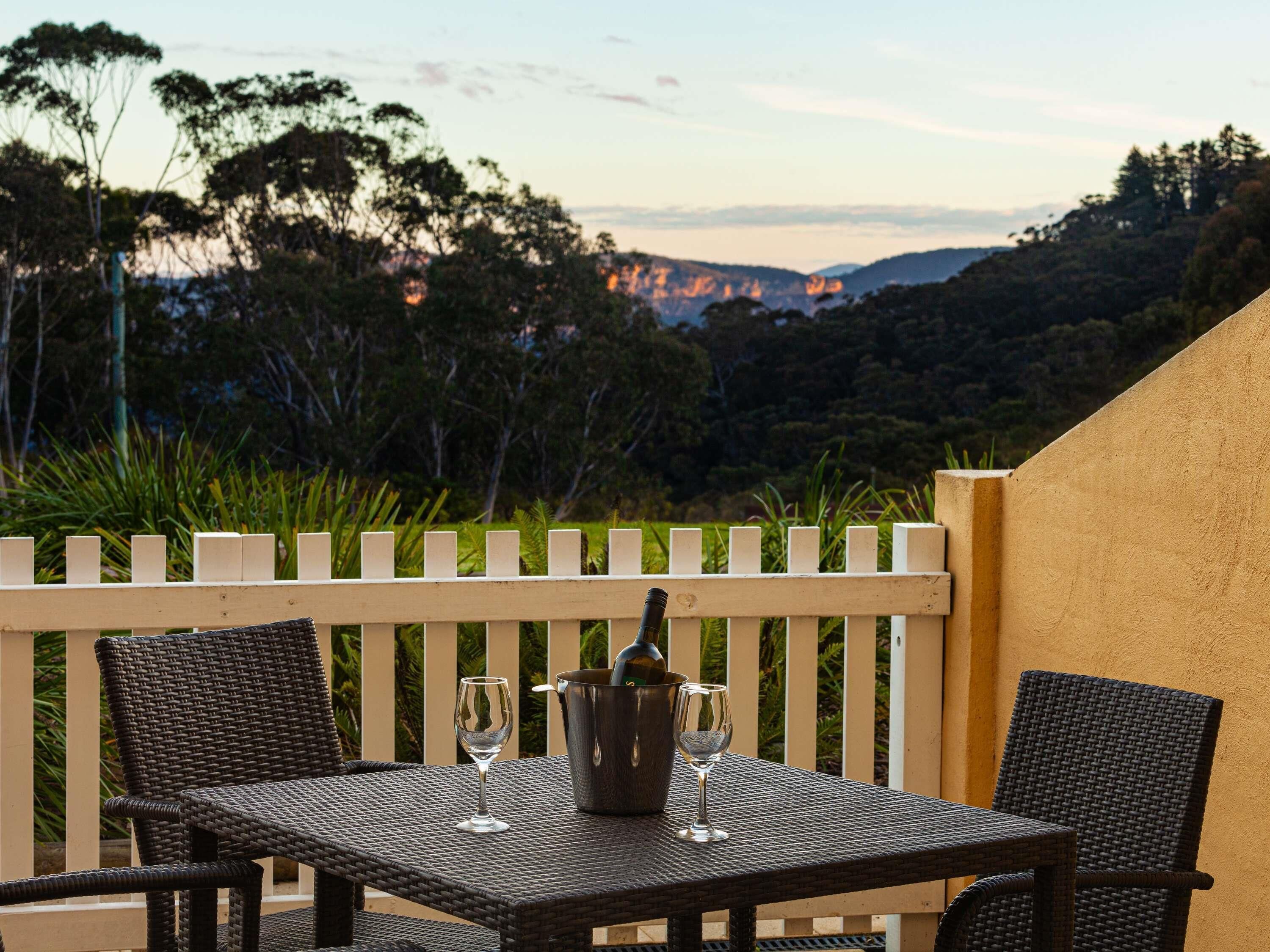 A table with two chairs and a wine bucket on a balcony with a view of trees and hills.