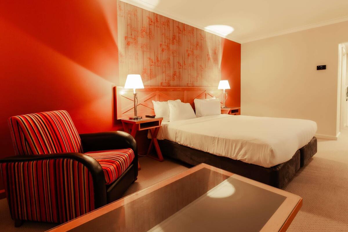 A hotel room with a large bed, two bedside tables with lamps, a striped armchair, and a glass coffee table.
