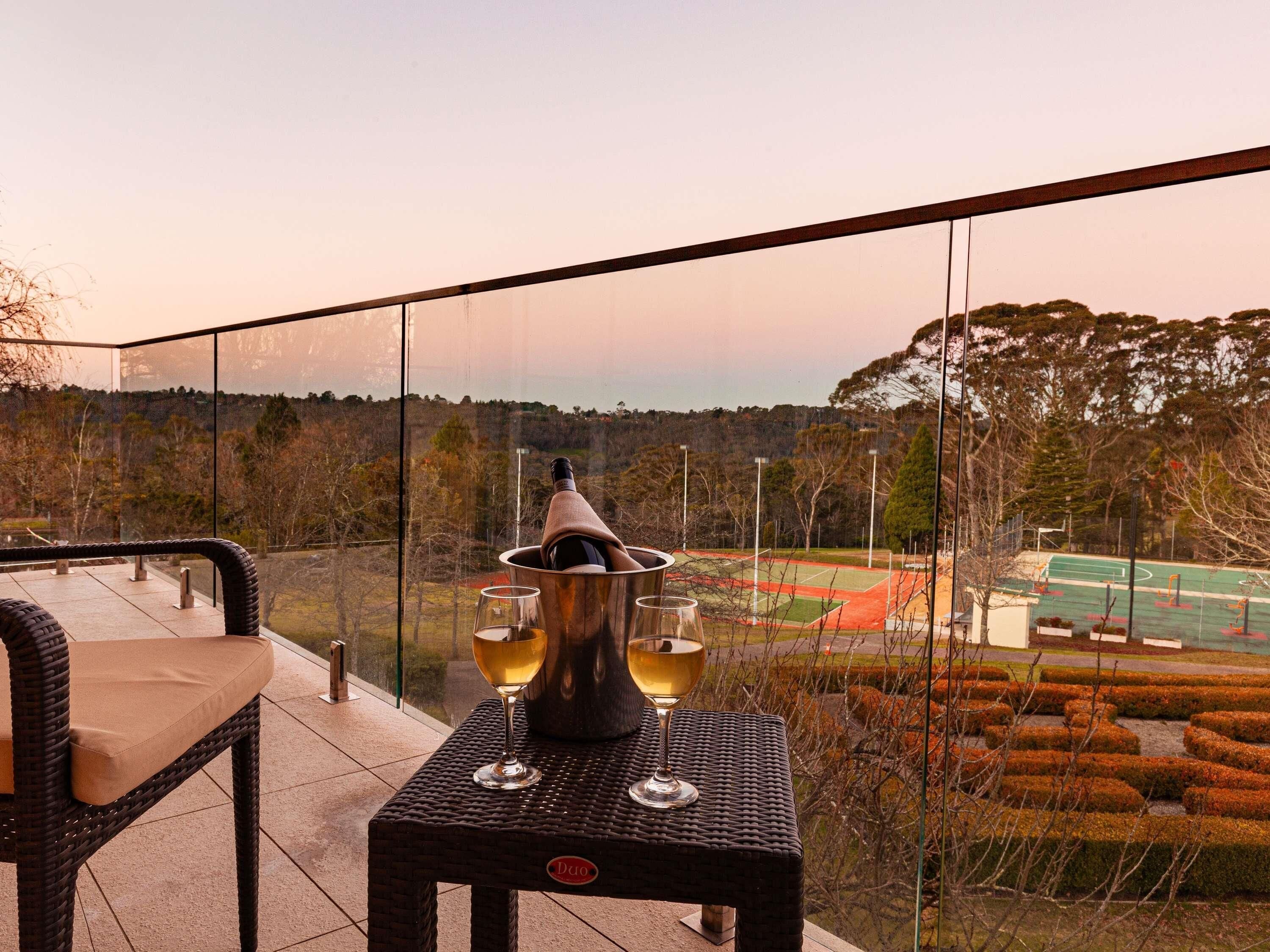 A balcony with wicker furniture, a table with a bottle and glasses, and a view of a sports field and trees.