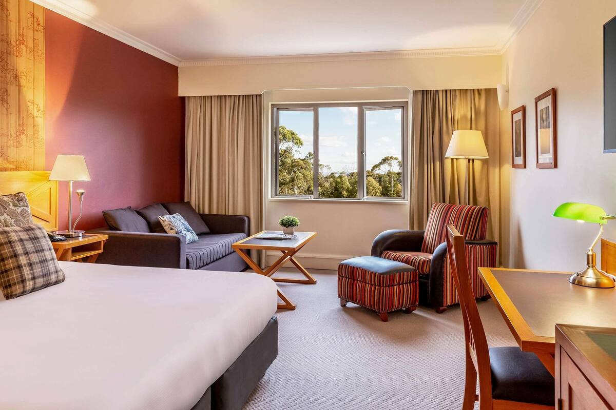 A hotel room with a bed, a sofa, a chair, a small table, and a window with a view of trees.