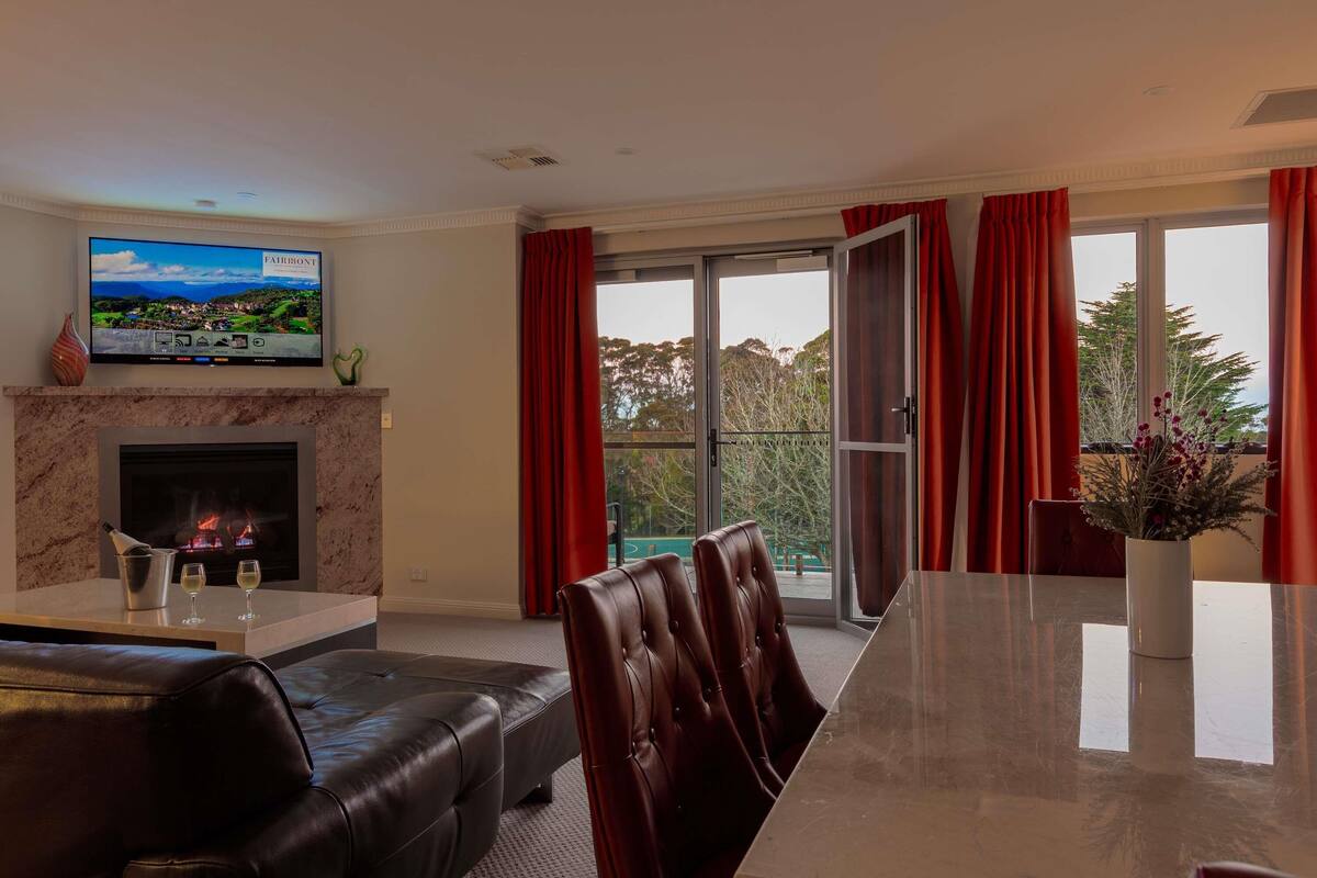 A modern living room with a fireplace, a flat-screen TV, a dining table, and red curtains.