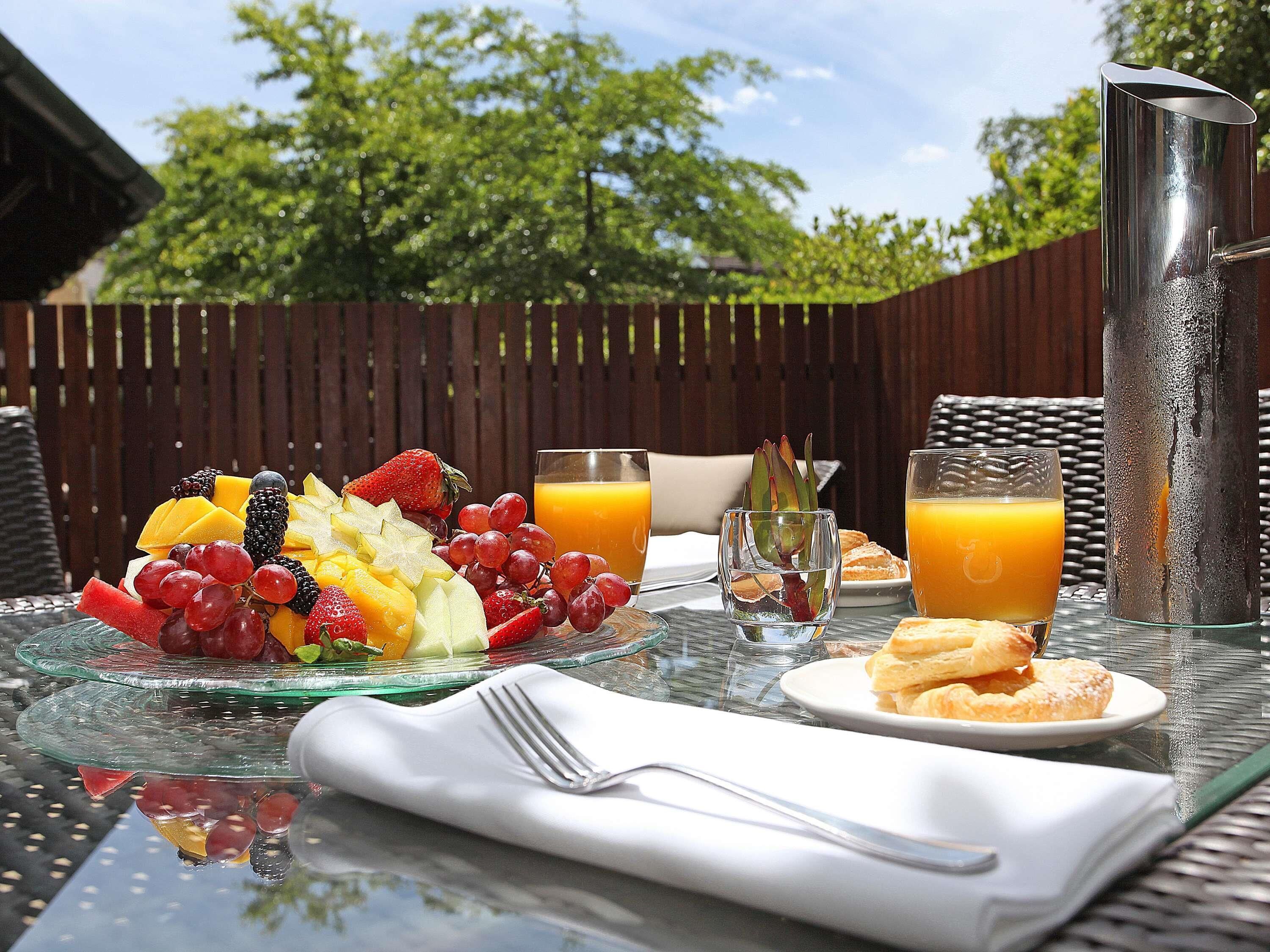 An outdoor breakfast setting with fruit, juice, and pastries on a glass table.