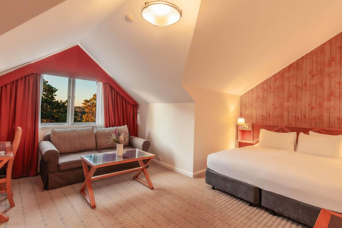 A hotel room with a bed, a sofa, a glass-top table, a window with curtains, and a ceiling light.
