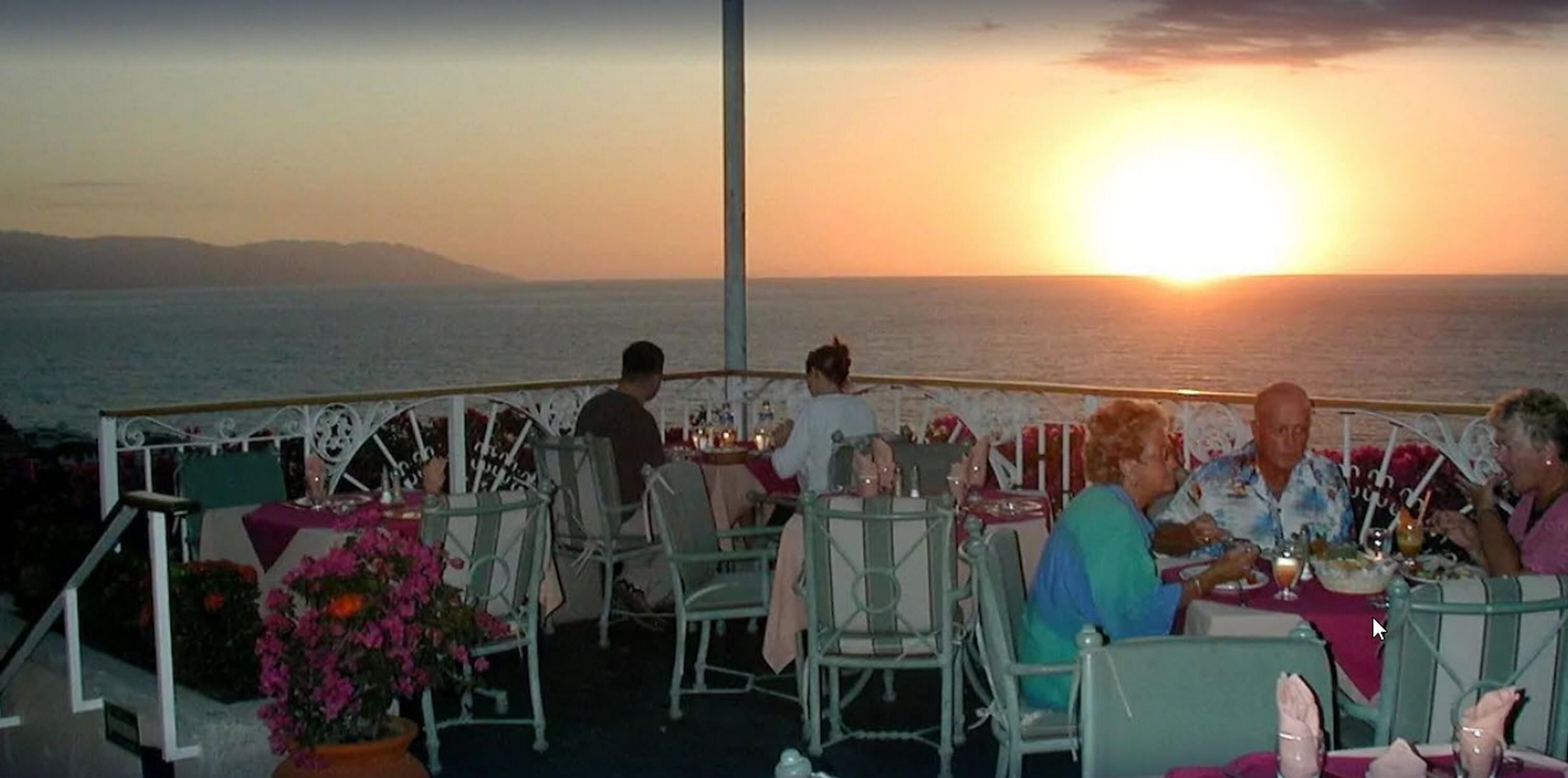 An outdoor dining area with tables and chairs, overlooking the sea at sunset.