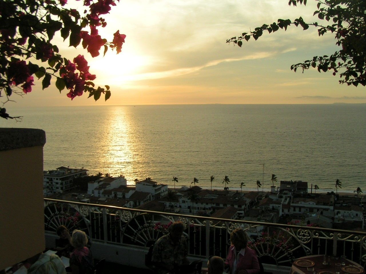 Un balcon donnant sur une vue imprenable sur la mer au coucher du soleil, avec des bâtiments en arrière-plan.