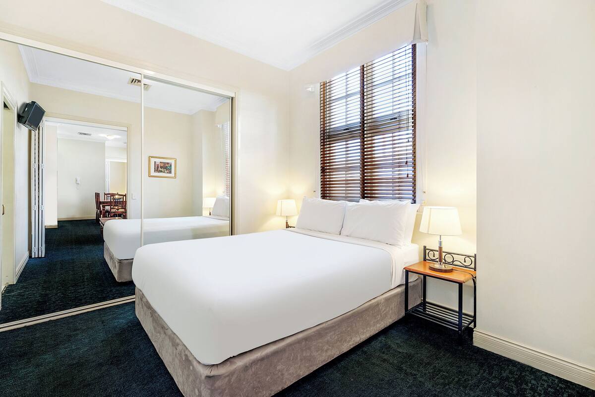 A hotel room with a large bed, a bedside table with a lamp, a mirror, and a window with blinds.