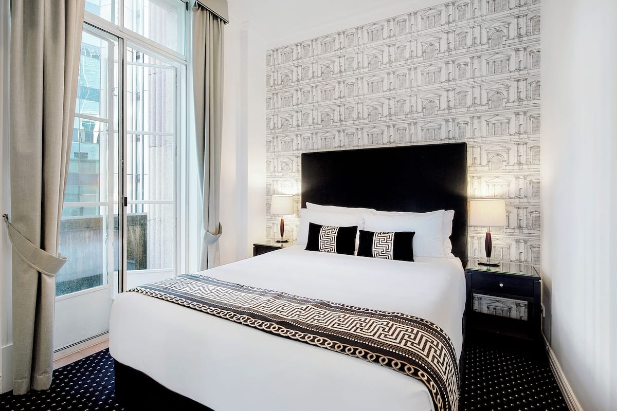 A hotel room with a large bed, a window with curtains, a patterned wall, and a nightstand with a lamp.